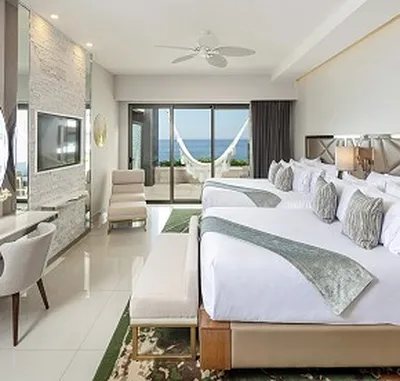 Modern hotel room with two large beds, a wall-mounted TV, and a balcony view of the ocean. A hammock is visible outside.