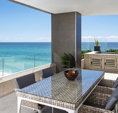 Outdoor patio with a wicker dining set on a balcony overlooking a clear blue ocean and sky, with potted plants and glass railing.