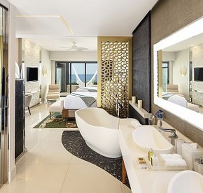 Luxurious hotel suite with a modern open-plan design, featuring a freestanding bathtub, double sinks, and a bed with a view of the ocean.
