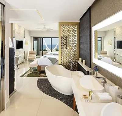 Luxurious hotel suite with a modern open-plan design, featuring a freestanding bathtub, double sinks, and a bed with a view of the ocean.