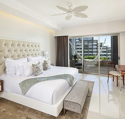 Bright hotel room with a large bed, white bedding, and decorative pillows. Sliding doors open to a balcony with a hammock and city view.