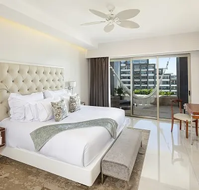 Bright hotel room with a large bed, white bedding, and decorative pillows. Sliding doors open to a balcony with a hammock and city view.