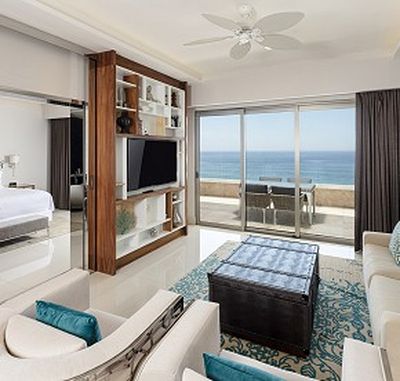 Modern living room with a sofa, coffee table, TV, and large glass doors opening to a balcony with ocean views. Bedroom visible through an open door.