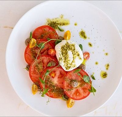 A plate of sliced tomatoes, cherry tomatoes, burrata cheese, greens, and pesto drizzle on a white plate.