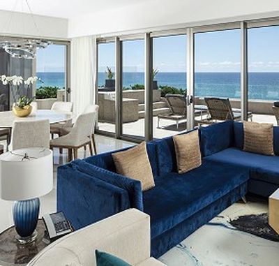 Luxurious living room with blue sofa, modern decor, and ocean view through large glass doors leading to a balcony with seating.