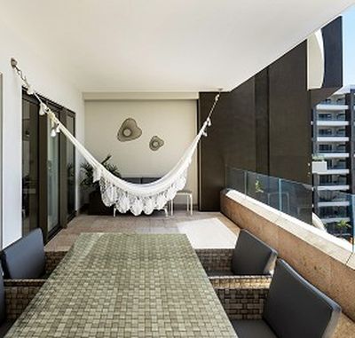 Modern balcony with a hammock, a mosaic-tiled table, and chairs. Glass railing offers a view of nearby buildings. Wall decor adds style.