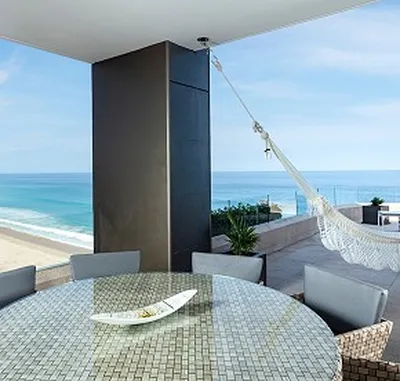 Rooftop patio with a glass table, gray chairs, and a hammock overlooking a sandy beach and ocean under a clear blue sky.