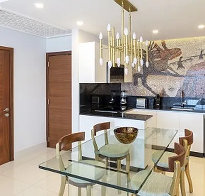 Modern kitchen with a glass dining table, wooden chairs, mosaic backsplash, and a gold chandelier. White cabinets and wooden doors complete the look.