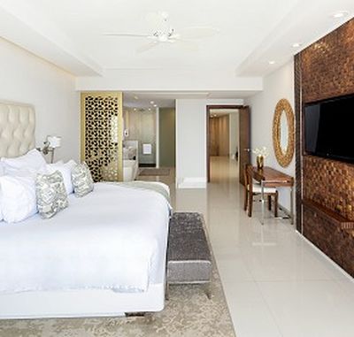 Modern hotel room with a large bed, decorative pillows, a wall-mounted TV, a wooden desk, and an open bathroom area with a partition.