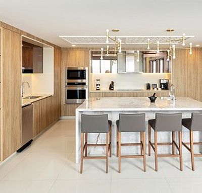 Modern kitchen with wooden cabinets, marble island, four gray bar stools, built-in appliances, and a stylish ceiling light fixture.