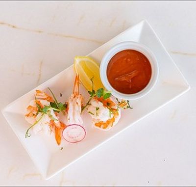 Shrimp cocktail with three shrimp, a lemon wedge, radish slice, and parsley on a rectangular plate, served with cocktail sauce in a small bowl.