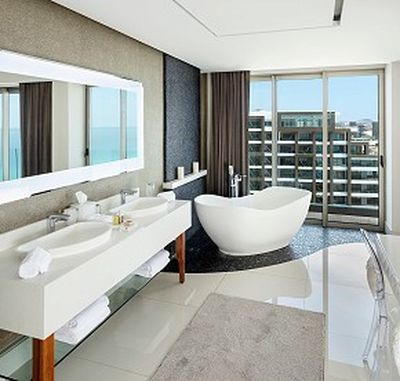 Modern bathroom with double sinks, large mirror, freestanding bathtub, floor-to-ceiling windows, and city view. Neutral tones and sleek design.