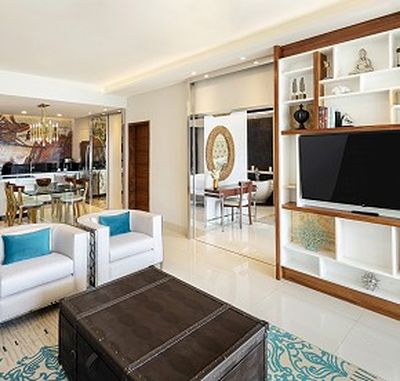 Modern living room with white sofas, turquoise cushions, a large TV on a wooden shelf, and a dining area in the background.