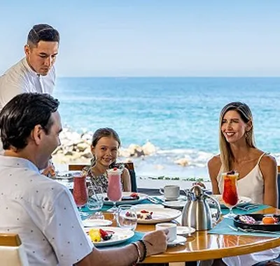 Family dining outdoors by the sea, served by a waiter. Smiling faces, colorful drinks, and a clear blue sky create a cheerful atmosphere.