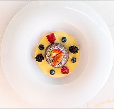A dessert with a chocolate cake topped with strawberries, surrounded by custard, blueberries, raspberries, and blackberries on a white plate.