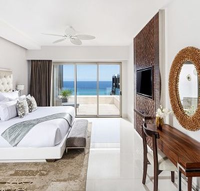 Modern hotel room with a king-size bed, ocean view balcony, wall-mounted TV, round mirror, and wooden desk with chair.