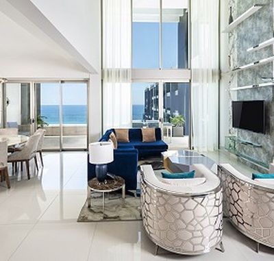 Modern living room with high ceilings, large windows, blue sofa, patterned chairs, and ocean view. Dining area visible on the left.