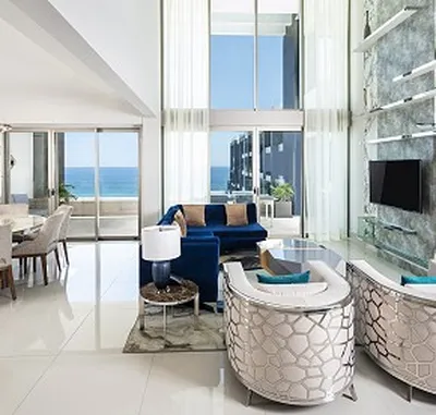 Modern living room with high ceilings, large windows, blue sofa, patterned chairs, and ocean view. Dining area visible on the left.