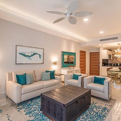 Modern living room with beige sofas, teal cushions, a wooden coffee table, ceiling fan, and wall art. Open view to dining area with chandelier.