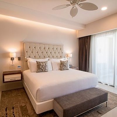 Elegant hotel room with a large bed, tufted headboard, bedside tables, lamps, a ceiling fan, and a bench. Light streaming through sheer curtains.
