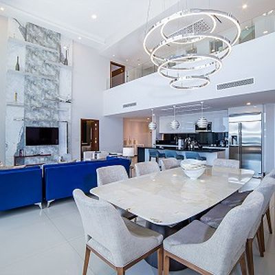 Modern open-plan living area with a marble dining table, blue sofas, and a sleek kitchen. Contemporary lighting and a double-height ceiling.