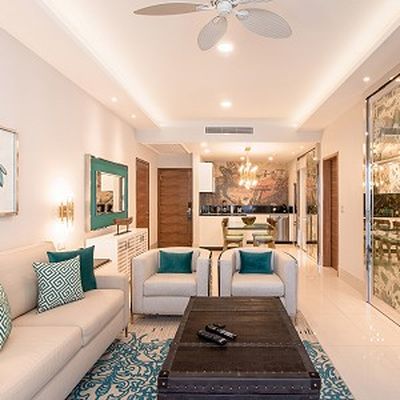 Modern living room with beige sofas, teal cushions, a black coffee table, and a ceiling fan. Open kitchen in the background with chandelier lighting.