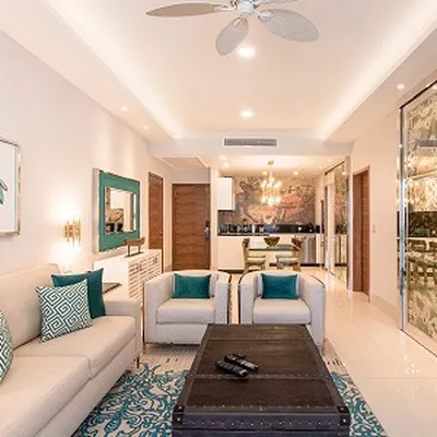 Modern living room with beige sofas, teal cushions, a black coffee table, and a ceiling fan. Open kitchen in the background with chandelier lighting.