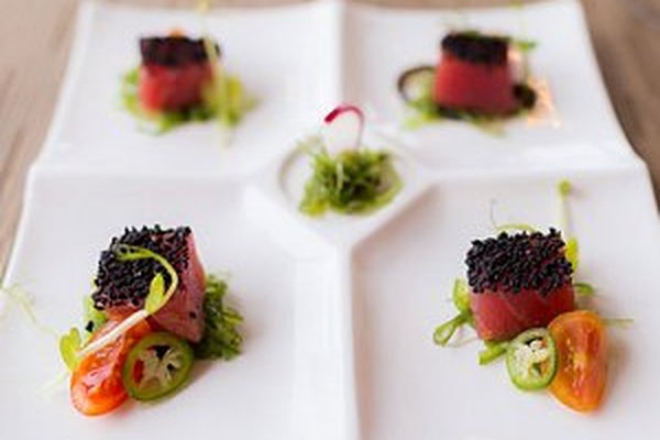 Gourmet dish with four square sections, each featuring seared tuna topped with black sesame seeds, garnished with greens and sliced vegetables.