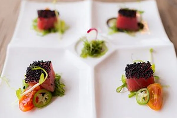 Gourmet dish with four square sections, each featuring seared tuna topped with black sesame seeds, garnished with greens and sliced vegetables.