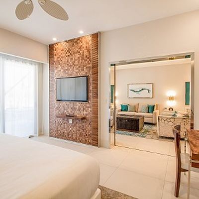 Modern hotel room with a wall-mounted TV, wooden accents, a bed, and a view into a living area with a sofa and artwork.