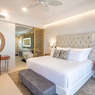 Elegant hotel room with a large bed, decorative pillows, a bench, a writing desk, and an open view into a modern bathroom.