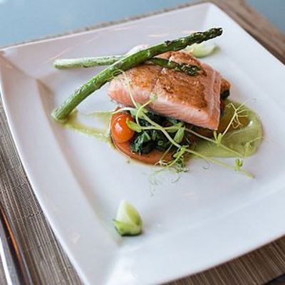 Grilled salmon fillet on a bed of spinach and cherry tomatoes, garnished with asparagus and pea shoots, on a white square plate.