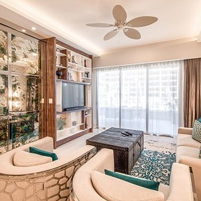 Bright living room with beige sofas, a wooden TV unit, large windows, a ceiling fan, and a patterned rug. Decor includes green accents and plants.