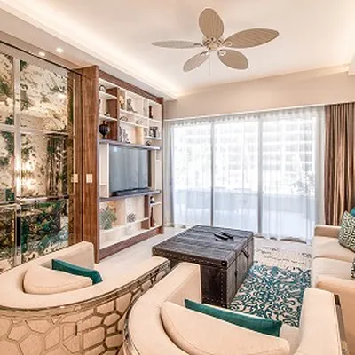Bright living room with beige sofas, a wooden TV unit, large windows, a ceiling fan, and a patterned rug. Decor includes green accents and plants.