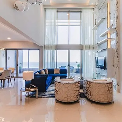 Spacious modern living room with high ceilings, large windows, blue sofa, two patterned chairs, and a wall-mounted TV. Dining area visible.