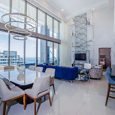 Modern living and dining area with large windows, ocean view, blue sofa, marble accent wall, and stylish lighting fixtures.