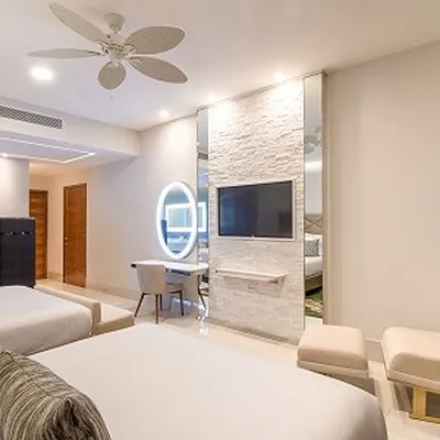 Modern hotel room with two beds, a wall-mounted TV, round mirror, and desk. Neutral tones and ceiling fan enhance the minimalist decor.