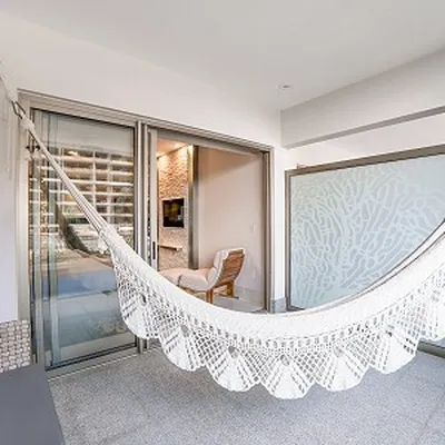A white hammock with decorative fringe hangs in a modern patio area, featuring sliding glass doors and a chair inside.