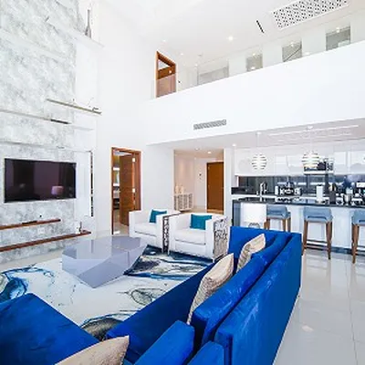 Modern living room with blue sofas, abstract rug, wall-mounted TV, and open kitchen. White walls and high ceiling create a spacious feel.