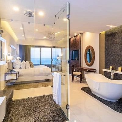 Luxurious hotel room with a glass-walled bathroom, freestanding tub, modern decor, and ocean view through large windows.