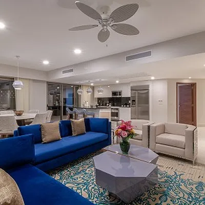 Modern living room with blue sofas, a geometric coffee table, and a ceiling fan. Open kitchen and dining area in the background.