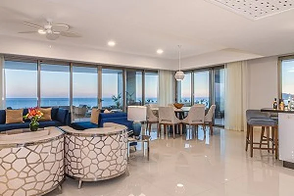 Spacious modern living room with ocean view, featuring blue sofas, patterned ottomans, dining area, and a bar with stools.