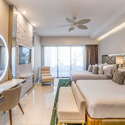 Modern hotel room with two large beds, a wall-mounted TV, a round mirror, and a seating area. Large window with sheer curtains.