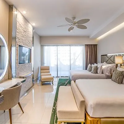 Modern hotel room with two large beds, a wall-mounted TV, a round mirror, and a seating area. Large window with sheer curtains.