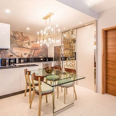 Modern kitchen with glass table, wooden chairs, mosaic wall art, chandelier, white cabinets, and stainless steel appliances.