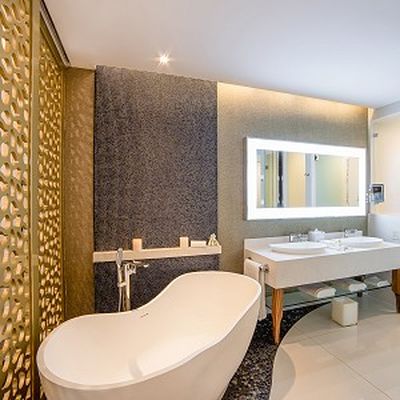 Modern bathroom with a freestanding white bathtub, decorative screen, large mirror with lights, and a double sink vanity on a tiled floor.