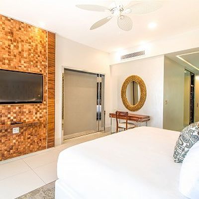 Modern hotel room with a large bed, wall-mounted TV, wooden accent wall, round mirror above a desk, and ceiling fan. Bright and inviting space.