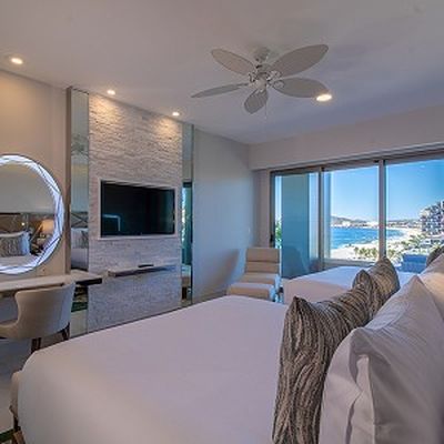 Modern hotel room with two beds, a wall-mounted TV, a round mirror, and a view of the ocean through large sliding glass doors.