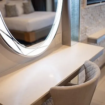 A modern vanity with a round mirror and a cushioned chair, set in a softly lit room with a bed in the background.