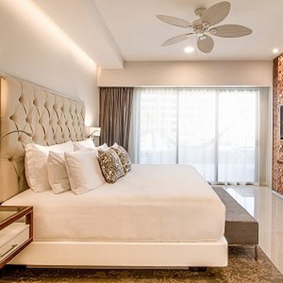Elegant hotel room with a large bed, tufted headboard, decorative pillows, ceiling fan, and floor-to-ceiling window with sheer curtains.